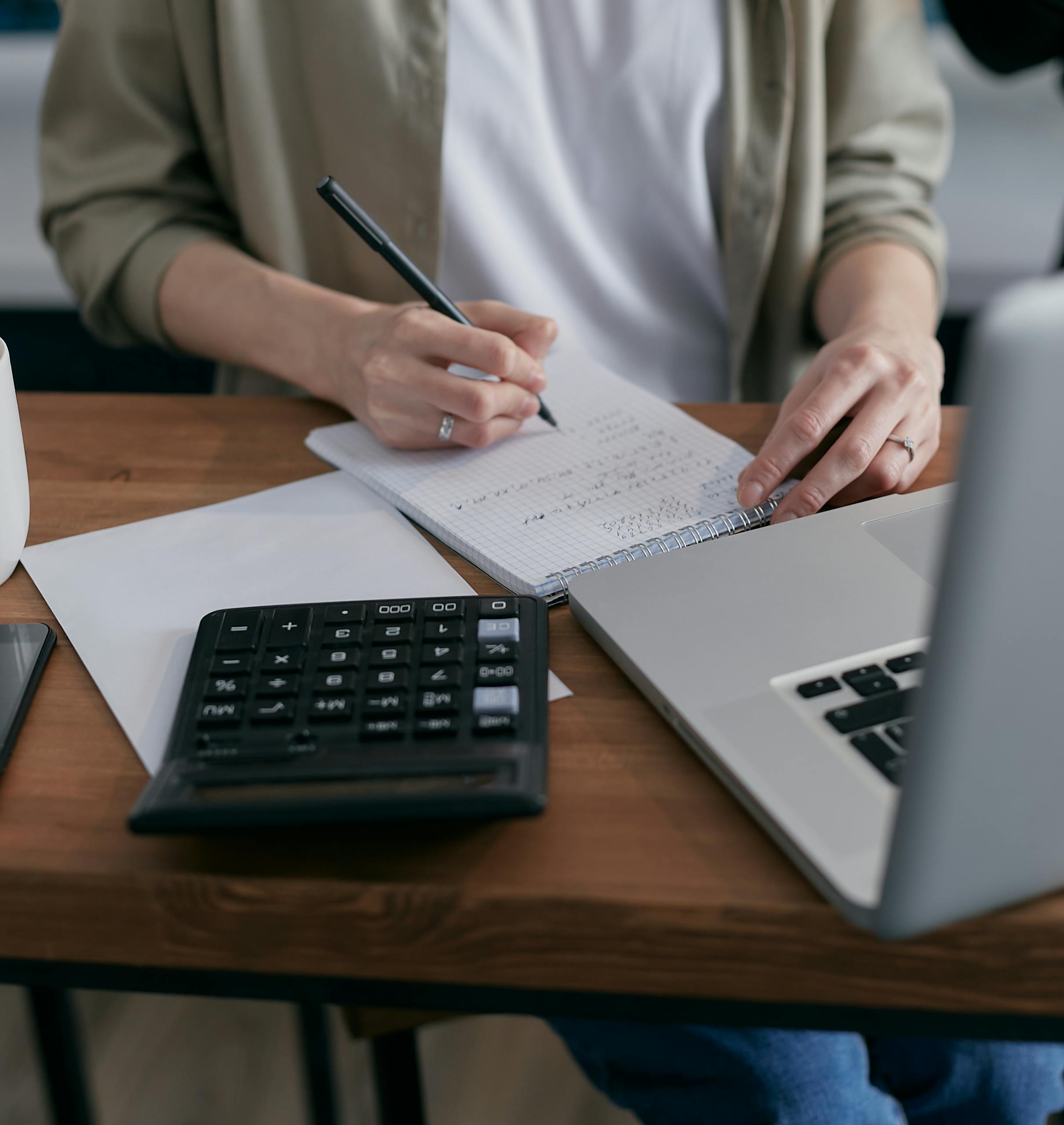 Resources 2 a woman writes financial calculations in a notebook using a calculator and laptop at a wooden desk. 6962993 scaled 1